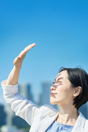 青空を背景に、手を掲げ眩しい日差しを遮る女性