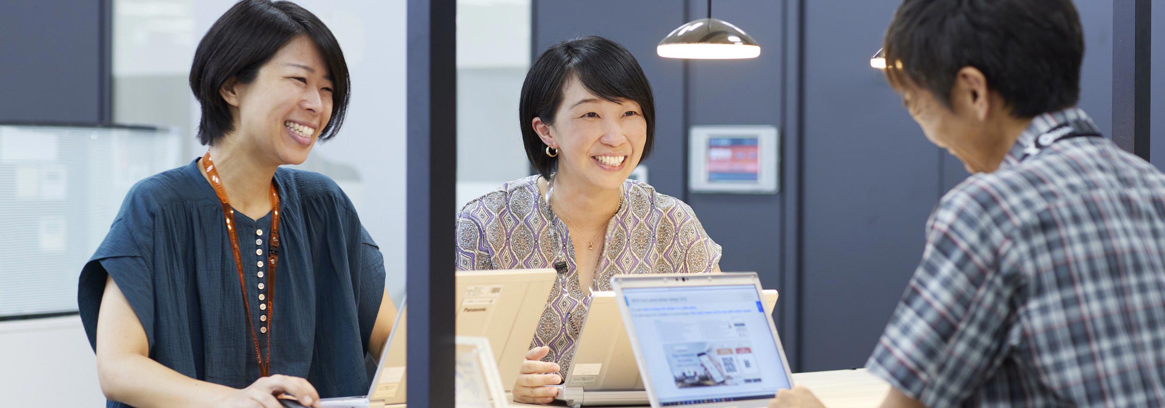ノートパソコンを手に同僚と談笑しながらミーティングをしている有本の画像